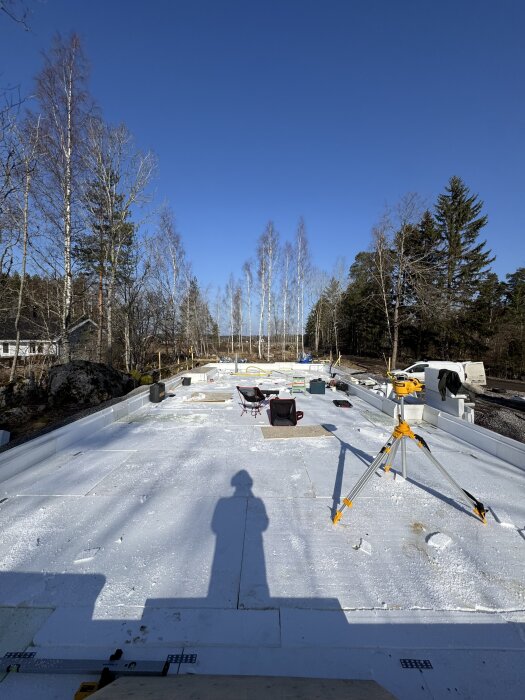 Byggarbetsplats med isolerat golv, trebent stativ i förgrunden, träd och klarblå himmel i bakgrunden.