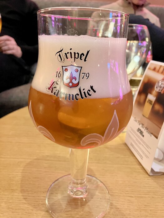 Ett glas Tripel Karmeliet öl med skumkrona på ett träbord, med suddiga personer i bakgrunden och en meny tavla vid sidan.