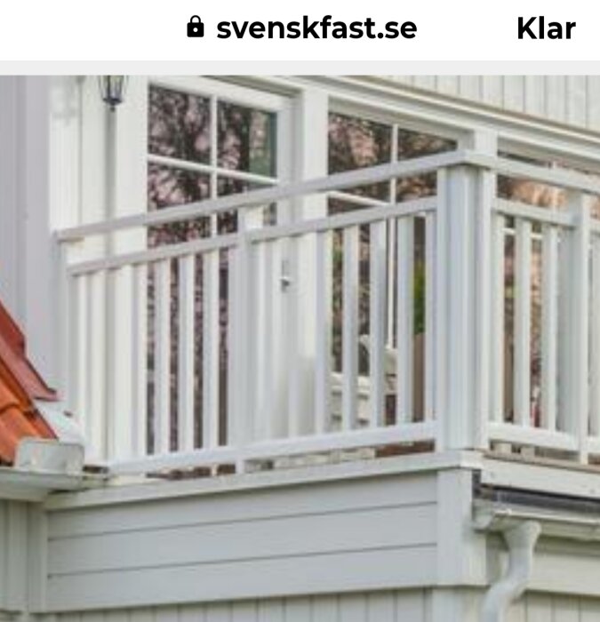 Balkong med vit trästaket, fönsterparti i bakgrunden och tak med röd takpanna synligt i hörnet.