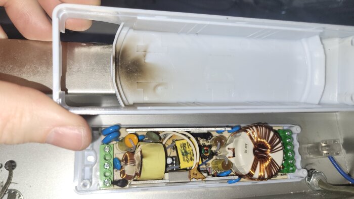 Närbild av en öppen elektrisk enhet med kretskort och kopparspolar, med en vit plastlock som uppvisar bränd rester.