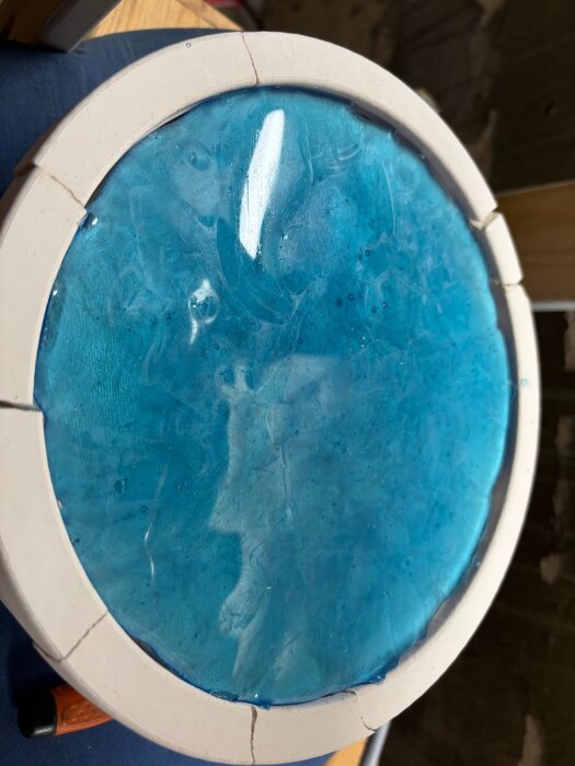 Blått glas i en rund form med ojämn yta och bubblor, omgivet av vit ram, placerat på en mörk bakgrund.