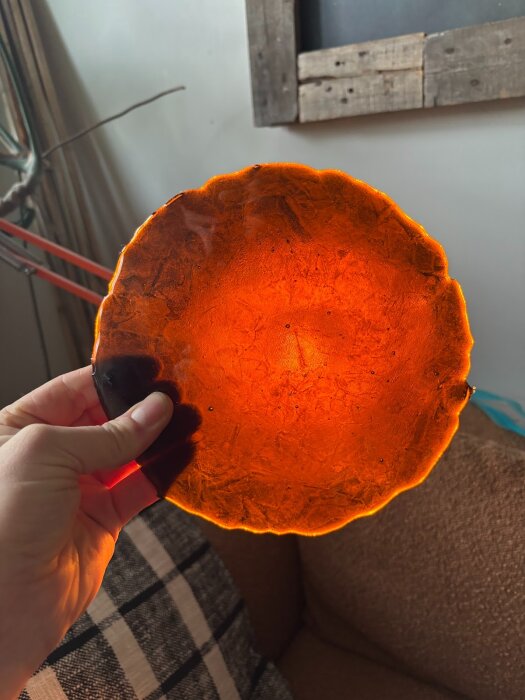 Rund, orange glasdisc hålls upp mot ljuset, med ojämn yta och svarta inneslutningar, visar tecken på värmebehandling och slumpingexperiment.