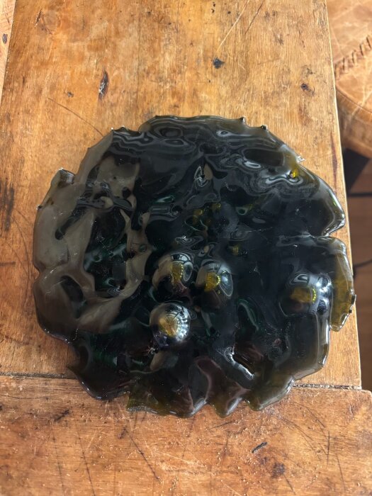 Smält och böljande glas med bubblor på träunderlag, glaset har en mörkgrönbrun färg och ojämn yta med synliga luftbubblor.