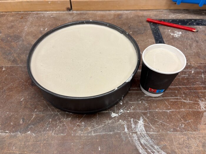 Rund gjutform för gipsstav och pappersmugg fylld med gipsblandning på träarbetsbänk, omgivna av verktygsspår och röd penna.
