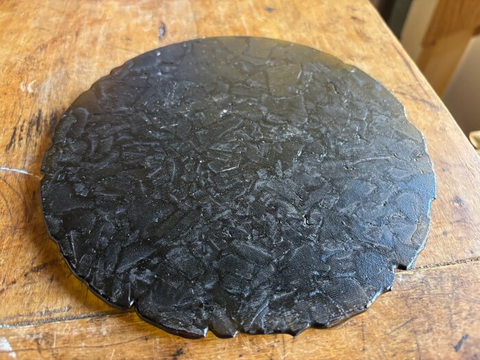 Svart, rund glasskiva med strukturerad yta på träbord. Resultat av experiment med glasbränning för att undersöka luftbubblors påverkan.