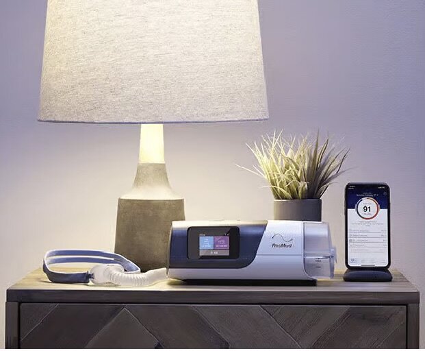 En CPAP-maskin, en smartphone med en sömnapp, och en bordslampa på ett nattduksbord.