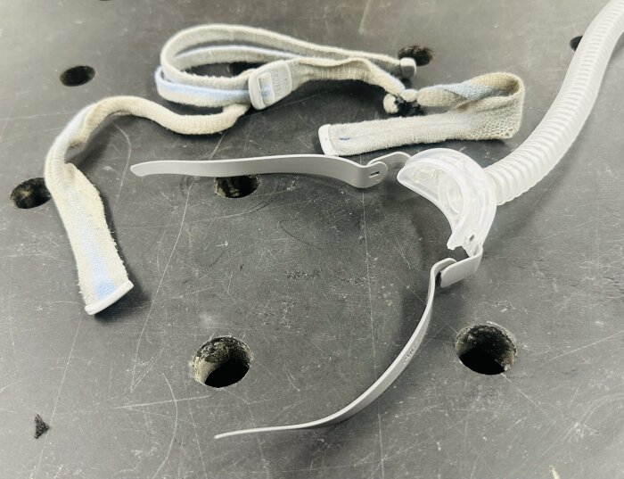 Delar av en näsmask för CPAP-maskin med remmar och en plastslang ligger på ett arbetsbord.