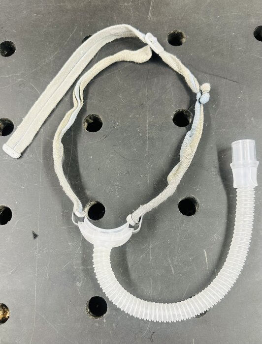 Justerad näsmask med elastiskt spännband och kardborrefästen, placerad på ett mörkt underlag med perforerade hål.