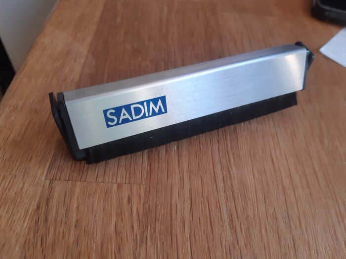 Kolfiber dammborste för grammofonskivor med texten "SADIM" på handtaget, avsedd att leda bort statisk elektricitet.