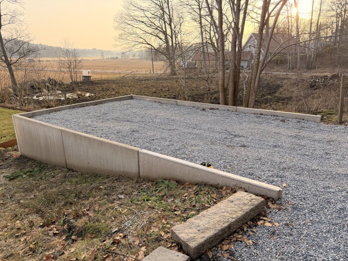 Grusyta med l-stöd inramar en plan markyta i sluttning, redo för carportbygge. Träd och hus i bakgrunden under solnedgång.