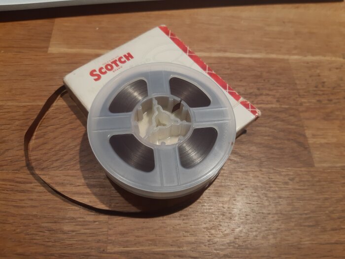 Rund bandrulle i plast med svart tejp liggande på en Scotch-förpackning på trägolv.
