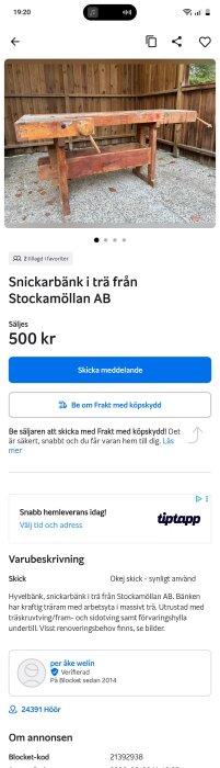 Snickarbänk i trä med kraftig arbetsyta och träskruvtving, står mot en trävägg på en grusyta. Bänken har visst renoveringsbehov.