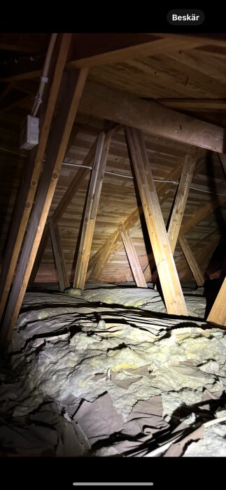 Loft med träbalkar och isolering syns på bilden; naturligt ljus lyser upp delar av trästrukturen och den fiberrika isoleringen.