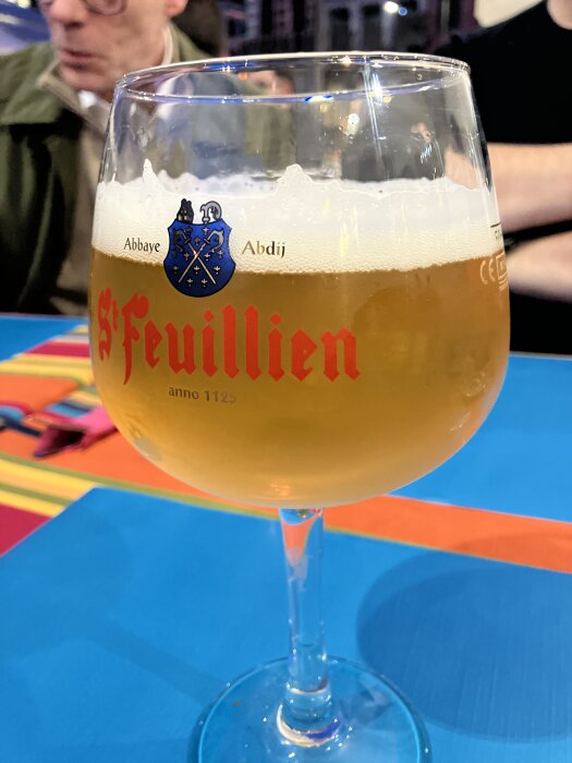 Ett glas St. Feuillien öl med skummande yta står på ett färgglatt bord.