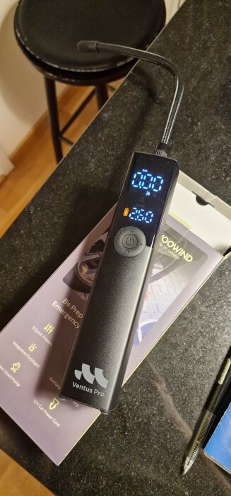 Digital luftpump Ventus Pro på en mörk bänkskiva, med en tryckdisplay och laddningskabel, bredvid en penna och förpackning.