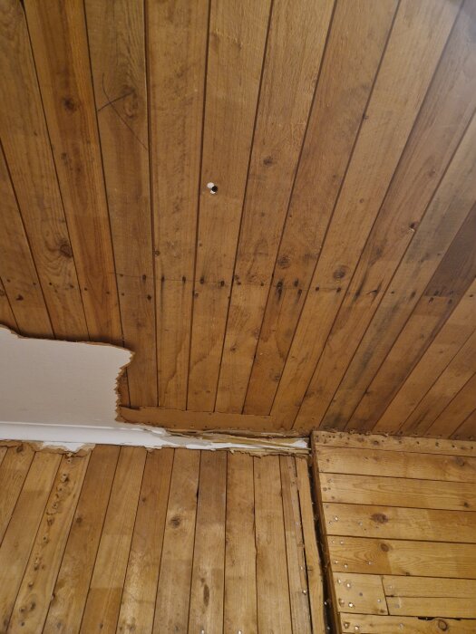 Träpanel på vägg och tak med delvis borttagen färg och avslöjad trästruktur. En skada eller renovation kan ha påbörjats.
