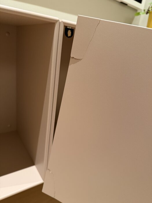 Sprucken MDF-lucka i ett skåp, närbild på spricka i och nära gångjärn; bakgrund är beige inredningsdetalj.