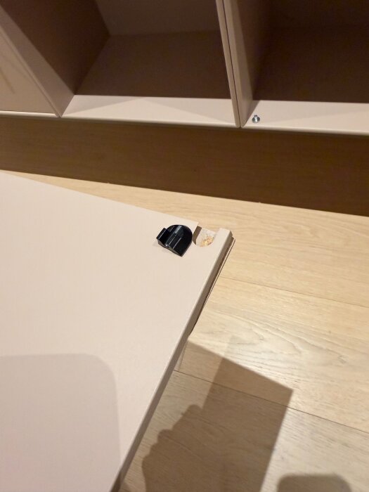 Sprucken MDF-lucka med synlig skada vid kanten samt ett litet löst gångjärn i plast placerat ovanpå luckan.