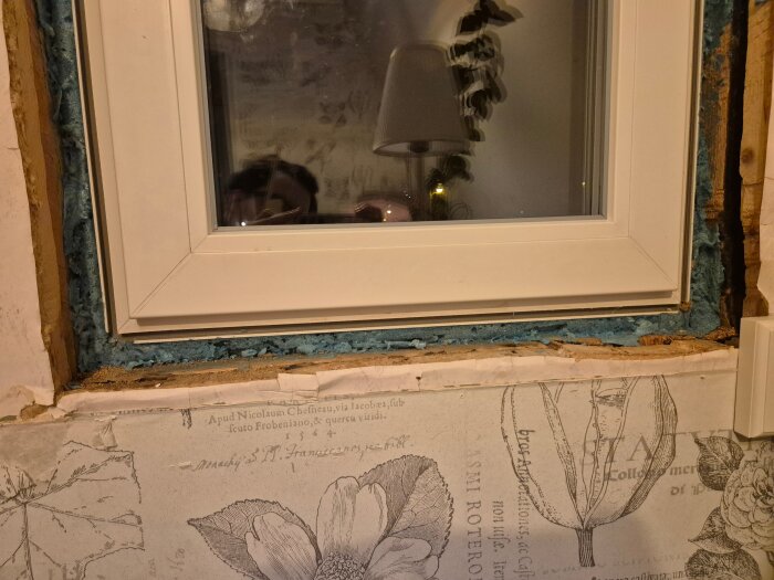 Närbild på ett fönster med blå isolering, exponerad karm och vägg med dekorativt tryck. Vattenskada och behov av nya fönstersmygar synlig.