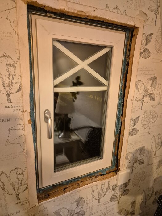 Fönster med vit karm och beslag, omgärdat av blottad vägg där fönstersmygen saknas. Tapet med blomstermotiv runt omkring.