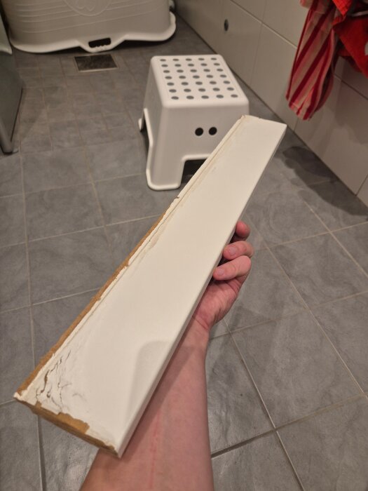 Vattenskadad fönstersmyg hålls i en hand i ett badrum med grått kakelgolv, vit pall i bakgrunden.