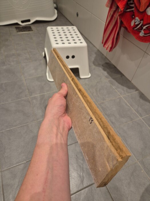 En hand håller en bit trälist som är del av en fönstersmyg, stående i ett kaklat badrum med en vit pall i bakgrunden.