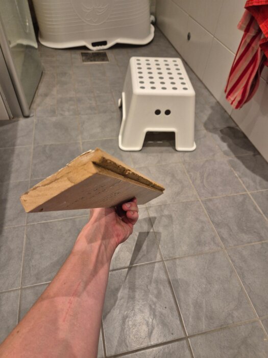 Hand håller i en träplanka över ett grått klinkergolv i ett badrum med en vit pall och en randig handduk i bakgrunden.