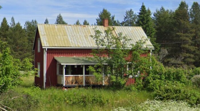 Ett äldre rött hus med vit veranda, omgärdat av träd och växtlighet, visar tecken på stort underhållsbehov.