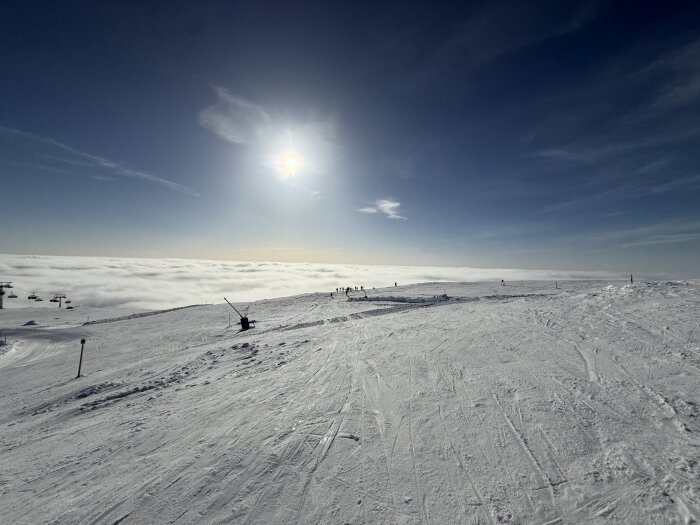 Snötäckt bergstopp under en klar himmel med solen som lyser och ett molntäcke i horisonten. Skidbacke syns i förgrunden.