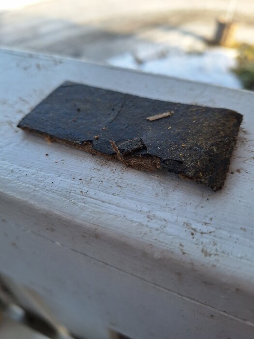 En bit av en svart papp/fibermatta med limrester, från ett hus byggt 1977, som misstänks innehålla asbest.