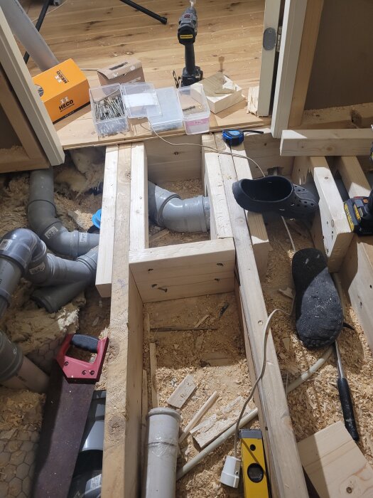 Byggarbetsplats med bjälklag, verktyg och avloppsrör. Träreglar och byggmaterial ligger omkring, inklusive en borrmaskin och en skruvmejsel.