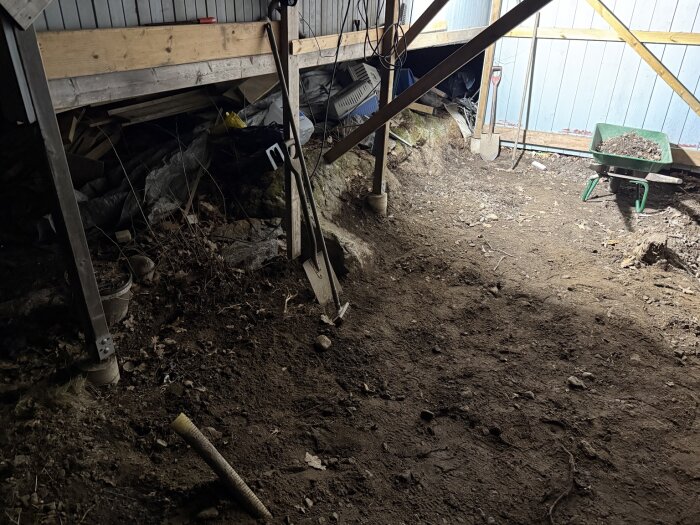 En pågående byggarbetsplats för ett garage med jordgolv, trästöd, berg i bakgrunden och en skottkärra med jord. Verktyg hänger på väggen.