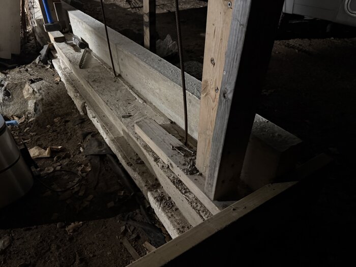 Form för gjutning av kantbalk med armeringsjärn och trästöd, del av garagebyggnation. Betong syns på marken bland grus och jord.