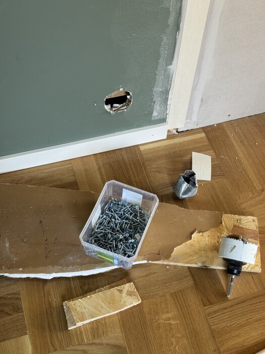 Hål i en grön vägg, omgiven av gipsskiva med dosborr och skruvar på golvet, under pågående renovering.