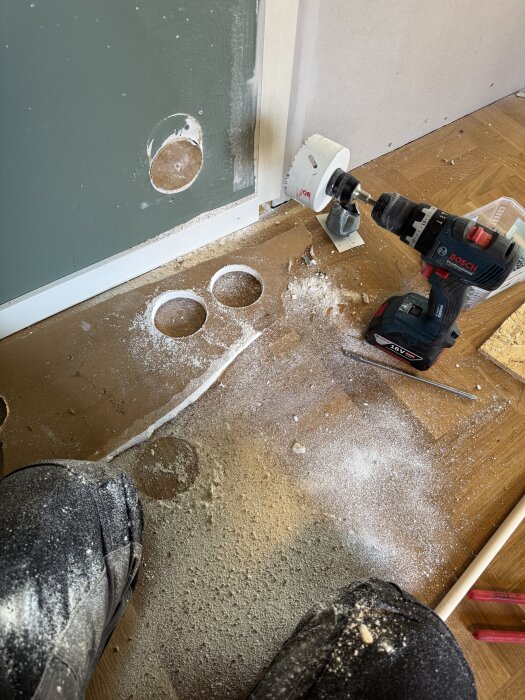 Borrmaskin med hålsåg borrar hål i en vägg under renovering, runt hål synligt, sågspån på golvet.