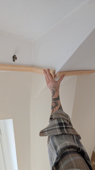 En person med tatuerad arm håller upp en trälist mot ett hörn i taket för att mäta eller installera den.
