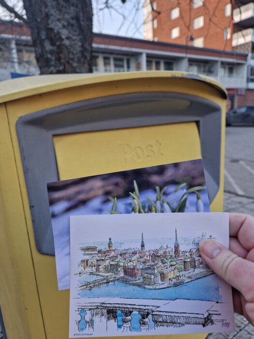 En hand håller vykort med Stockholm-motiv och snödroppar framför en gul brevlåda utomhus.