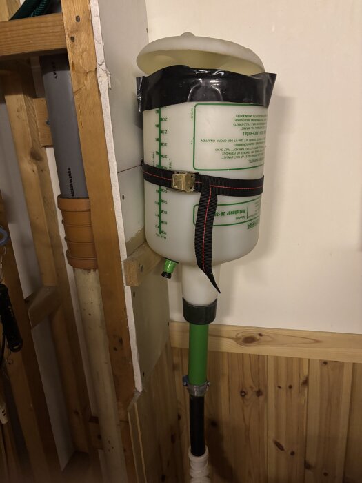 Vit behållare med vatten upp till hälften, fastsatt med rem, del av hydroforanläggning i trä- och rörmiljö.