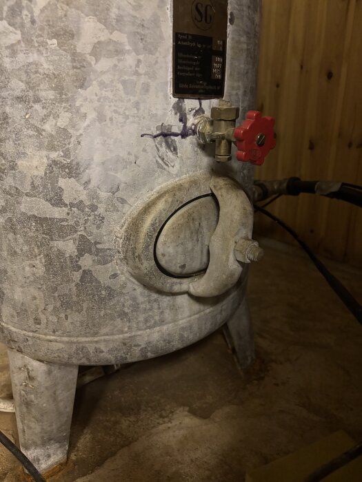 Botten av en hydrofor med en ventil och en röd ratt märkt "SG", kopplad till rör i en träpanelklädd källare.