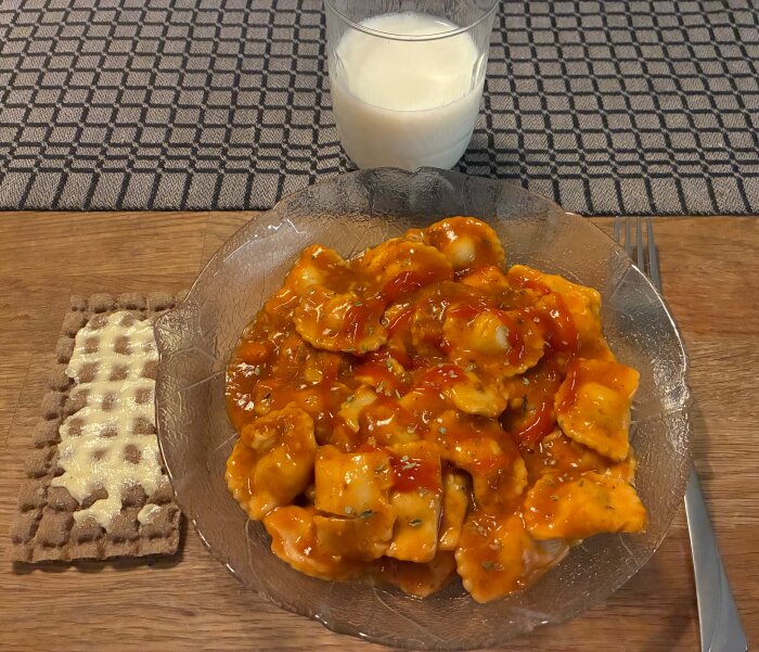 En tallrik ravioli med tomatsås, ett glas mjölk och en knäckebrödsskiva med smör på ett träbord med bordstablett.