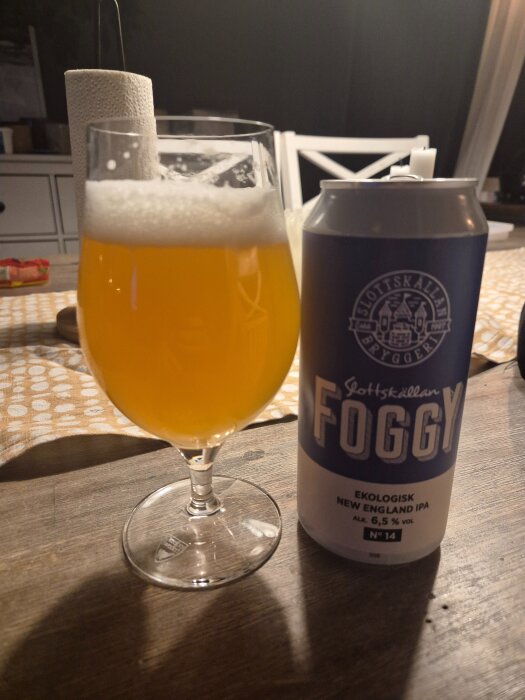 Ett glas med disig, gul IPA-öl och en burk märkt "Slottskällan Foggy Ekologisk New England IPA" på ett bord.
