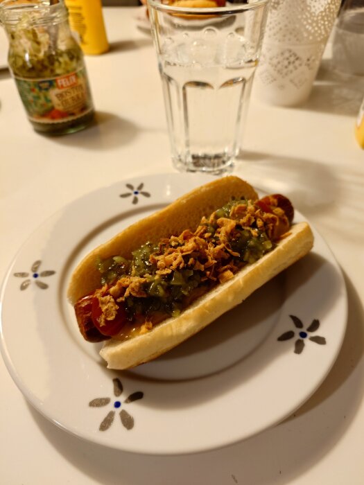 Grillad korv med bröd, toppad med gurkmajonnäs och rostad lök på en vit tallrik. Glas med dryck och syltburk i bakgrunden.