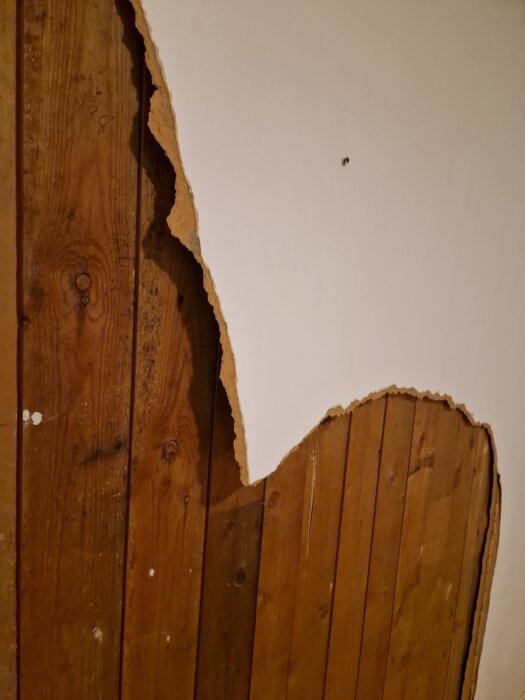 Riven masonitskiva på vägg med vit målad yta och träpanel bakom.