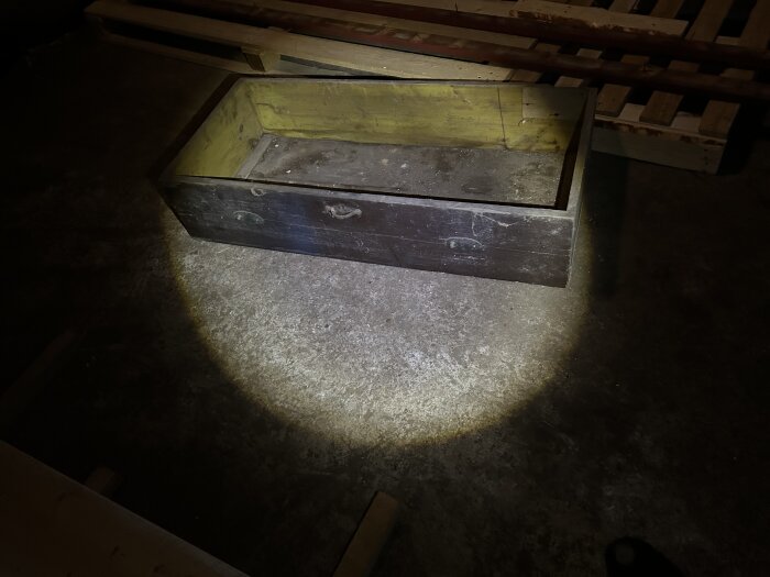 Gammal byrålåda i trä, gul inuti, står på betonggolv i ett uthus, belyst av ficklampa.