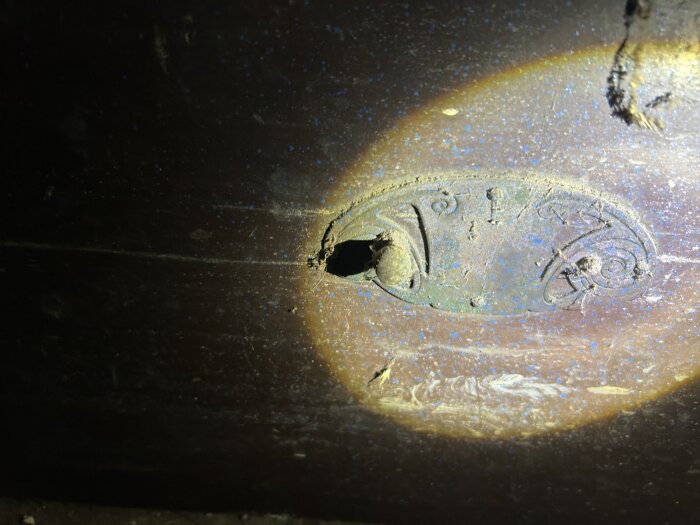 En gammal träbyrålåda i ett uthus, med en liten metallplakett eller märke inpräglat på framsidan, belyses av en ficklampa.