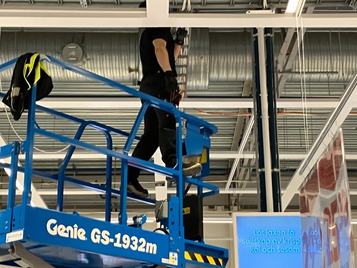 En person balanserar på räcket av en saxlift vid taket i en butik.