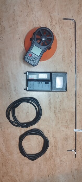 Verktyg för luftflödesmätning: en digital anemometer, kompaktmätare, två svarta kablar och en L-formad sond på ett träbord.