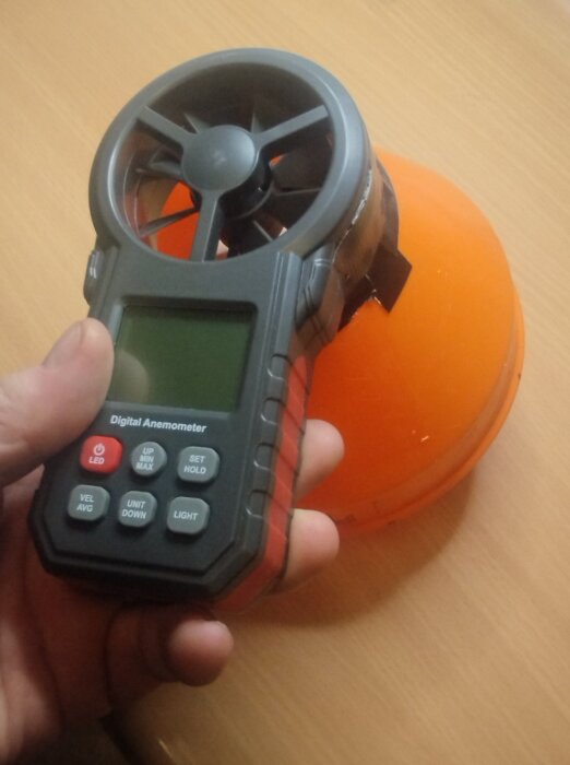 Hand håller en digital anemometer nära en orange kon på ett bord.