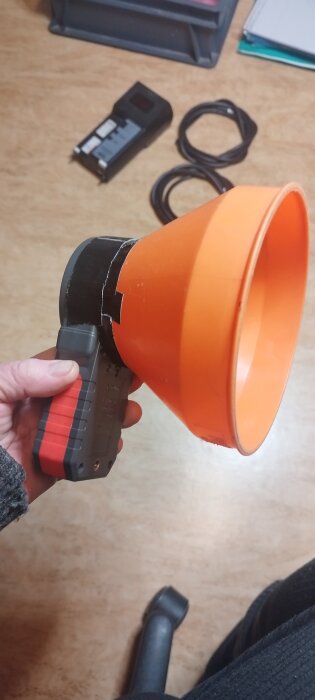 Hand håller en luftflödesmätare med orange tratt och digital display, placerad på ett bord med mätkablar och mätverktyg i bakgrunden.