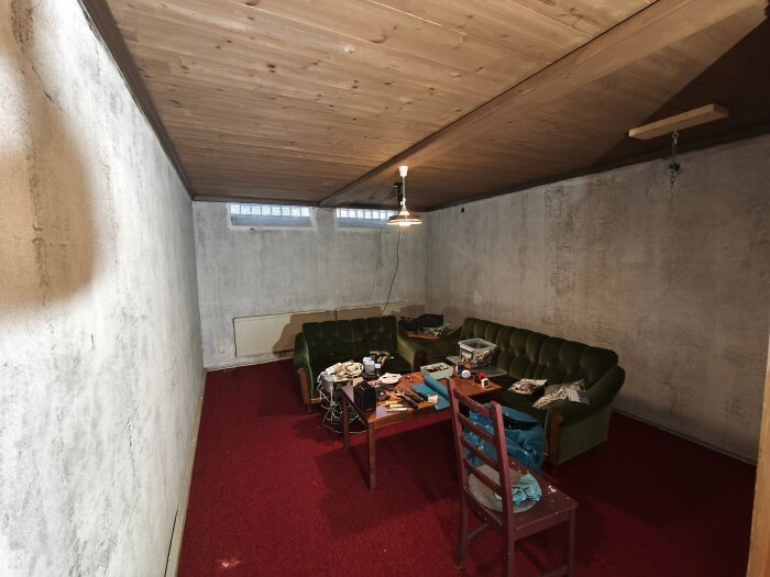 Gillestuga med slitna betongväggar, gröna soffor, rött golv och träpaneltak, med en lampa hängande från taket. Bord fullt med verktyg och material.